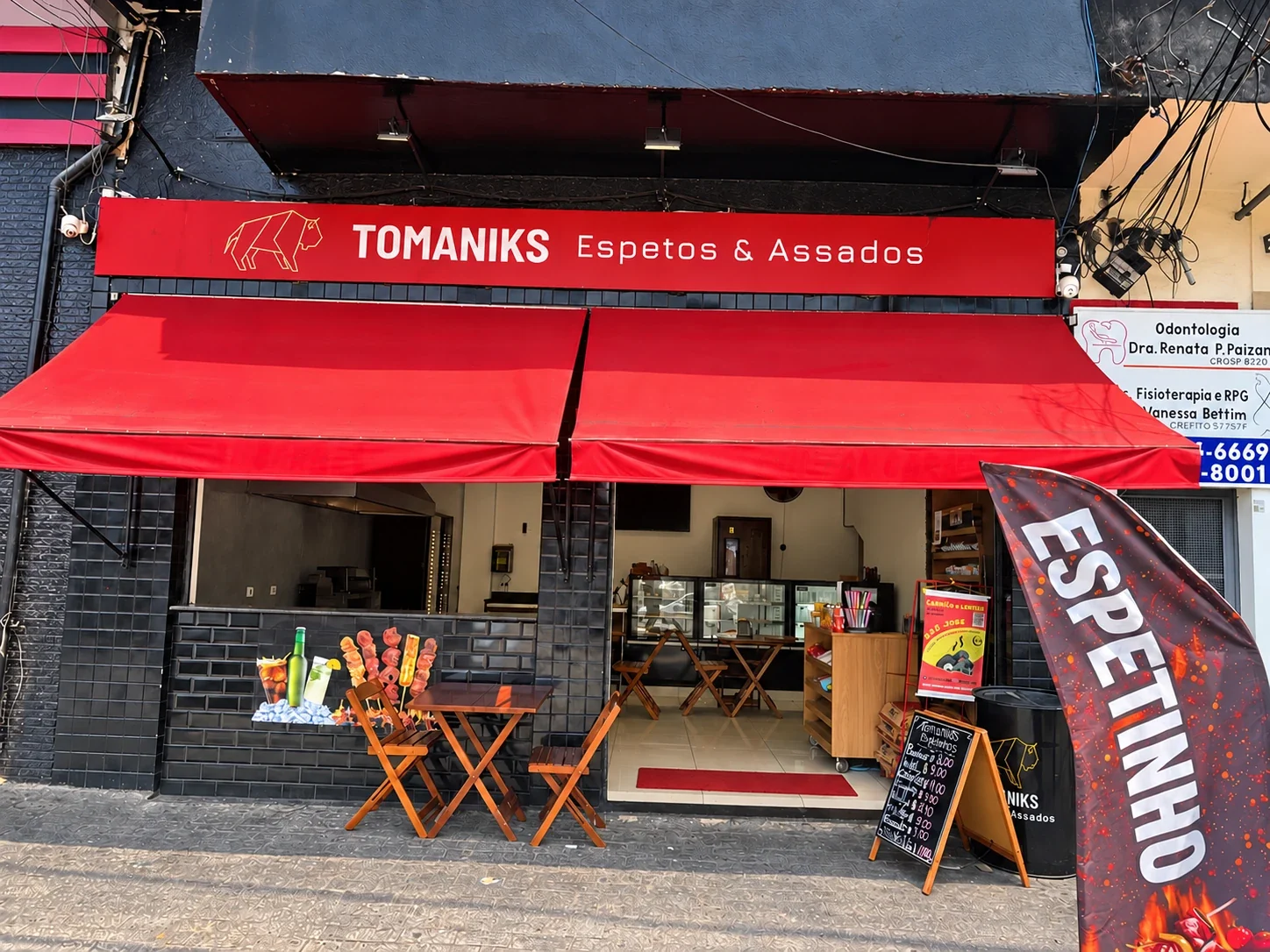 Fachada da Tomaniks Espetos & Assados, com toldos vermelhos e entrada sem pessoas.